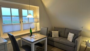 A cozy room with a table, a chair, a sofa, a window with blinds, a lamp, and a bottle of wine. - Söl'ring Hüs SO/19 - Söl'ring Hüs (Sylt)