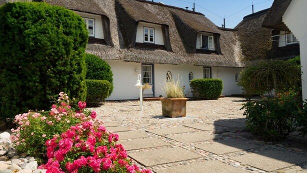 Una casa tradicional con techo de paja y un patio de piedra, rodeada de setos cuidados y plantas con flores.