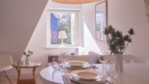 A dining area with a table set for two, a vase with flowers, and a window with a view of a building. - Söl`ring Hüs S0/29 - Söl'ring Hüs (Sylt)