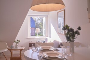 Et spiseområde med et bord dækket til to personer, en vase med blomster og et vindue med udsigt til en bygning.