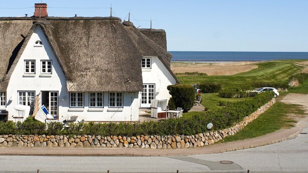 A thatched-roof house with a white facade, a stone wall, and a view of the sea. - Söl`ring Hüs S0/29 - Söl'ring Hüs (Sylt)