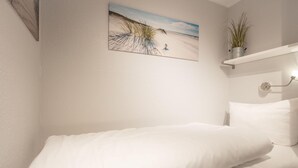 A bedroom with a single bed, a wall-mounted shelf with a plant, and a beach-themed painting. - SO/10 Söl`ring Hüs Raantem Lek - Söl'ring Hüs (Sylt)