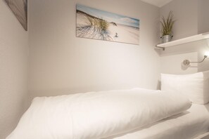 Une chambre avec un lit simple, une étagère fixée au mur ornée d’une plante, et un tableau sur le thème de la plage.