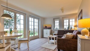 A spacious living room with a wooden floor, a brown sofa, a white dining table with chairs, and large windows offering a view of the outdoors. - SO/10 Söl`ring Hüs Raantem Lek - Söl'ring Hüs (Sylt)
