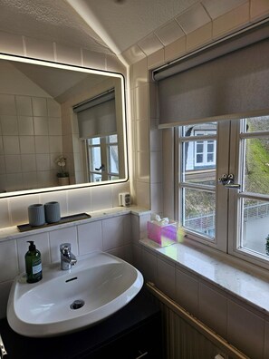A bathroom with a white sink, a mirror with a light, and a window with a view of the outside. - Söl`ring Hüs "Wattkieker" SO/23 - Söl'ring Hüs (Sylt)