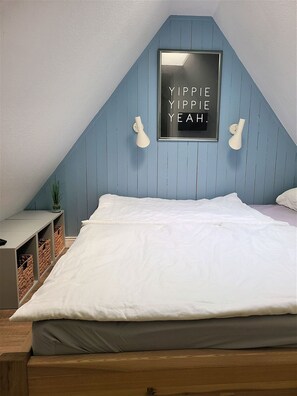 A bedroom with a sloped ceiling, a bed with white bedding, two wall-mounted lamps, and a framed sign on the wall. - Söl`ring Hüs "Wattkieker" SO/23 - Söl'ring Hüs (Sylt)