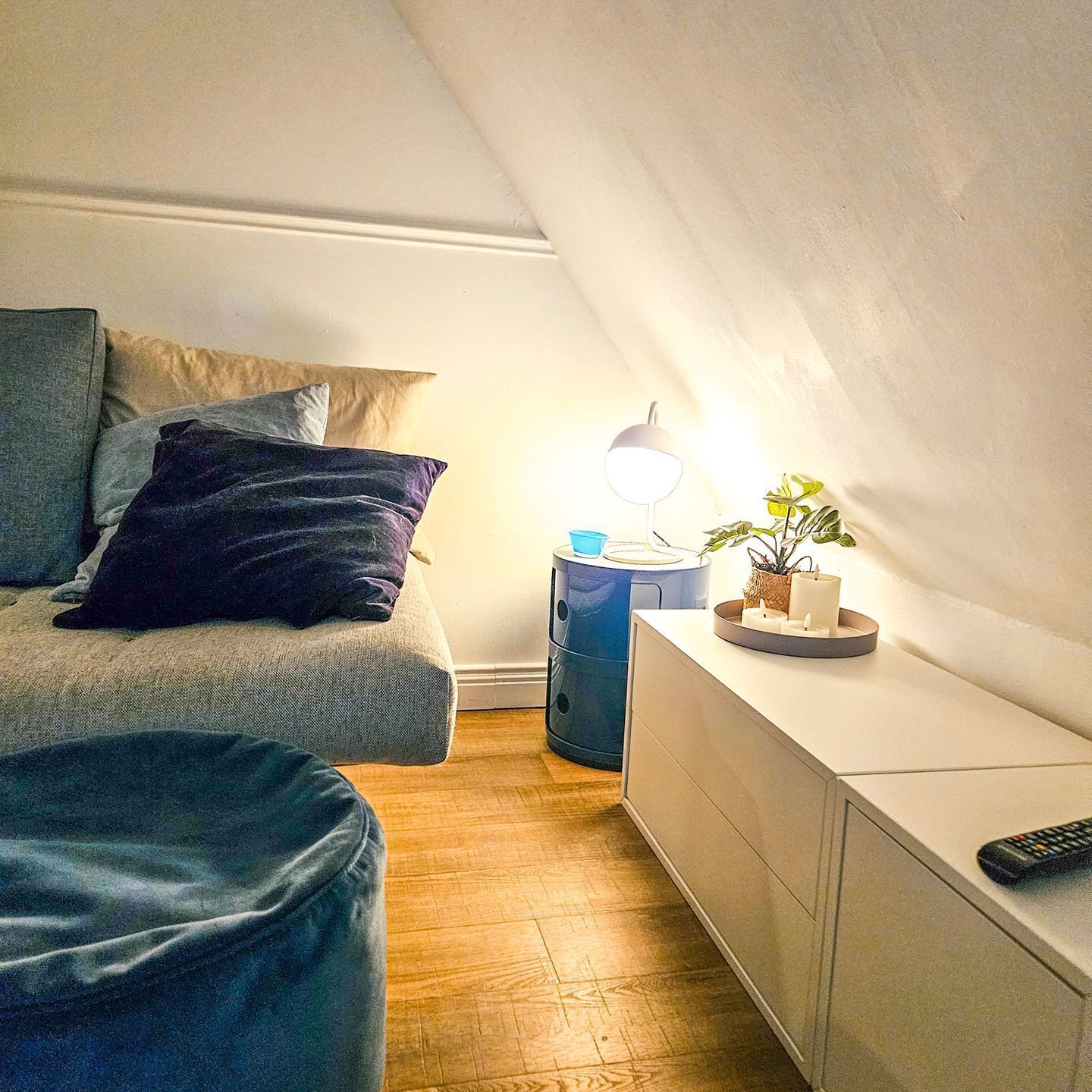 Una habitación acogedora con una cama, una mesita de noche con lámpara, una planta pequeña y un mando a distancia sobre un mueble blanco.