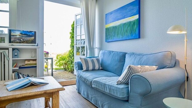 A living room with a blue sofa, a small table, a bookshelf, and a painting on the wall. - Söl`ring Hüs "Meerblick" SO/06 - Söl'ring Hüs (Sylt)