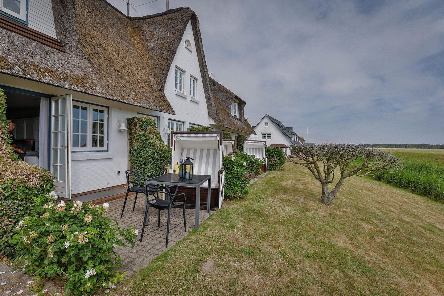 A row of white houses with thatched roofs, a patio with a table and chairs, and a small tree in the foreground.