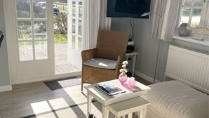 A cozy living room with a sofa, a chair, a small table with a magazine, a TV, and a view of the outdoors through large windows.