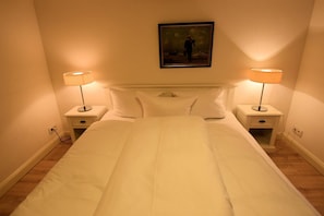Una cama bien hecha con ropa blanca, dos mesitas de noche con lámparas y un cuadro enmarcado en la pared.
