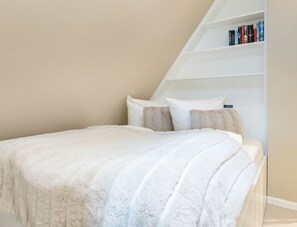 A bedroom with a white bed, a white bookshelf, and a beige wall. - Raantem Hüs App. 09 - Raantem Hus (Sylt)