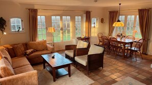 Un salon chaleureux avec un canapé, des fauteuils, une table basse et une table à manger avec des chaises. De grandes fenêtres sont ornées de rideaux, une lampe est présente, ainsi qu’une petite table sur laquelle sont posés divers objets.