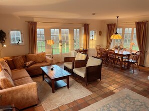 A cozy living room with a sofa, chairs, a coffee table, and a dining table with chairs. There are large windows with curtains, a lamp, and a small table with items on it. - Hellas Hüs - Hellas Hüs ST/48 (Sylt)