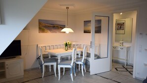 A dining area with a white table and chairs, a television on a stand, and artwork on the walls. - Strandstraße 26 - ST/26 Im Dünen Wai 07 (Sylt)