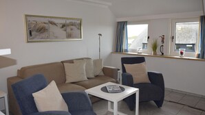 A living room with a beige sofa, two blue armchairs, a white coffee table, and a large painting above the sofa. - Strandstraße 26 - ST/26 Im Dünen Wai 07 (Sylt)