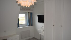 A bedroom with a bed, a window with curtains, a TV mounted on the wall, and a door. - Strandstraße 26 - ST/26 Im Dünen Wai 07 (Sylt)