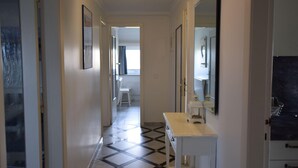 A hallway with a tiled floor, a white bench, and a small table with a mirror. There are framed pictures on the walls and a door on the left. - Strandstraße 26 - ST/26 Im Dünen Wai 07 (Sylt)