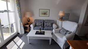 A living room with a white sofa, striped armchair, and a coffee table. There are two lamps, a framed picture on the wall, and a window with curtains. - House Wattblick App. 02 - Wattblick (Sylt)