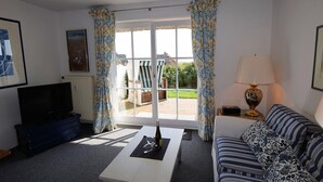 A living room with a TV, a sofa, a coffee table with a bottle and glasses, and a view of the outside. - House Wattblick App. 02 - Wattblick (Sylt)