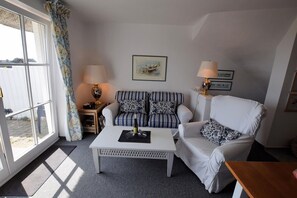 Ein Wohnzimmer mit einem weißen Sofa, einem gestreiften Sessel und einem Couchtisch. Zwei Lampen, ein gerahmtes Bild an der Wand und ein Fenster mit Vorhängen sind ebenfalls zu sehen.