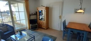 A room with a glass door leading to a balcony, a wooden cabinet, a dining table with chairs, and a hanging light fixture. - House Wattblick App. 03 - Wattblick (Sylt)