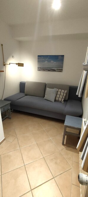 A living room with a grey sofa, a small table, a wall-mounted lamp, and a painting of a beach scene. - Wattblick House, Apt. 03 - Wattblick (Sylt)