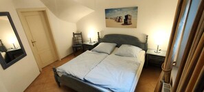Une chambre à coucher avec un lit, deux tables de chevet avec des lampes, une chaise, un tableau au mur et une porte.
