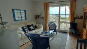 Een woonkamer met een bank, een glazen bijzettafeltje en een fauteuil. Er staat een televisie op een dressoir en een raam met gordijnen.