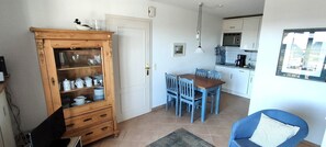 A compact kitchen with white cabinets, a dining table with blue chairs, and a wooden cabinet with dishes. - House Wattblick App. 03 - Wattblick (Sylt)