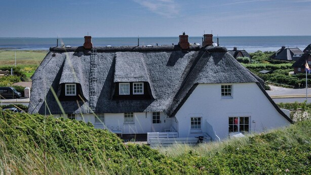 A traditional thatched-roof house with a white facade, situated on a grassy hill overlooking a coastal area. - House Wattblick App. 03 - Wattblick (Sylt)
