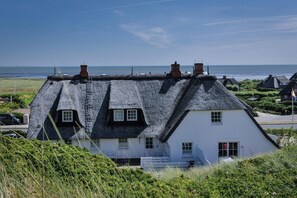 A traditional thatched-roof house with a white facade, situated on a grassy hill overlooking a coastal area. - House Wattblick App. 03 - Wattblick (Sylt)