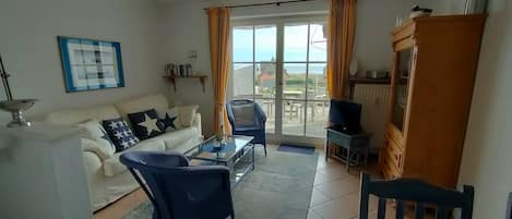 A living room with a sofa, a glass coffee table, and a chair. There is a television on a stand and a window with curtains.