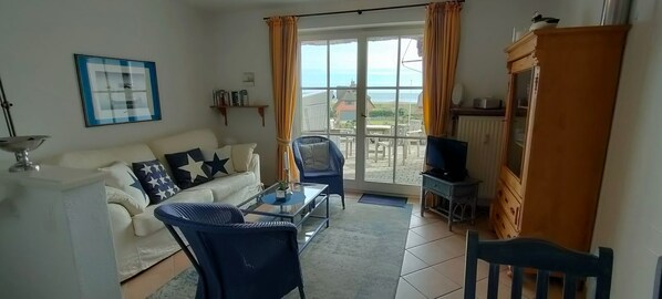 A living room with a sofa, a glass coffee table, and a chair. There is a television on a stand and a window with curtains. - House Wattblick App. 03 - Wattblick (Sylt)