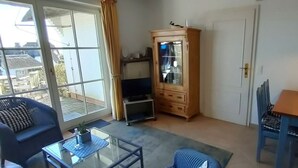 Een kamer met een glazen deur die leidt naar een balkon, een houten kast, een eettafel met stoelen en een hanglamp.