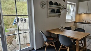 Ein Essbereich mit Holztisch und -stühlen, eine Küche mit weißen Schränken und Blick auf eine grüne Außenanlage durch eine Glastür.