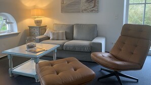 Ein Wohnzimmer mit grauem Sofa, einem braunen Ledersessel, einem kleinen Tisch, einer Lampe und einem Bild mit Strandmotiv.
