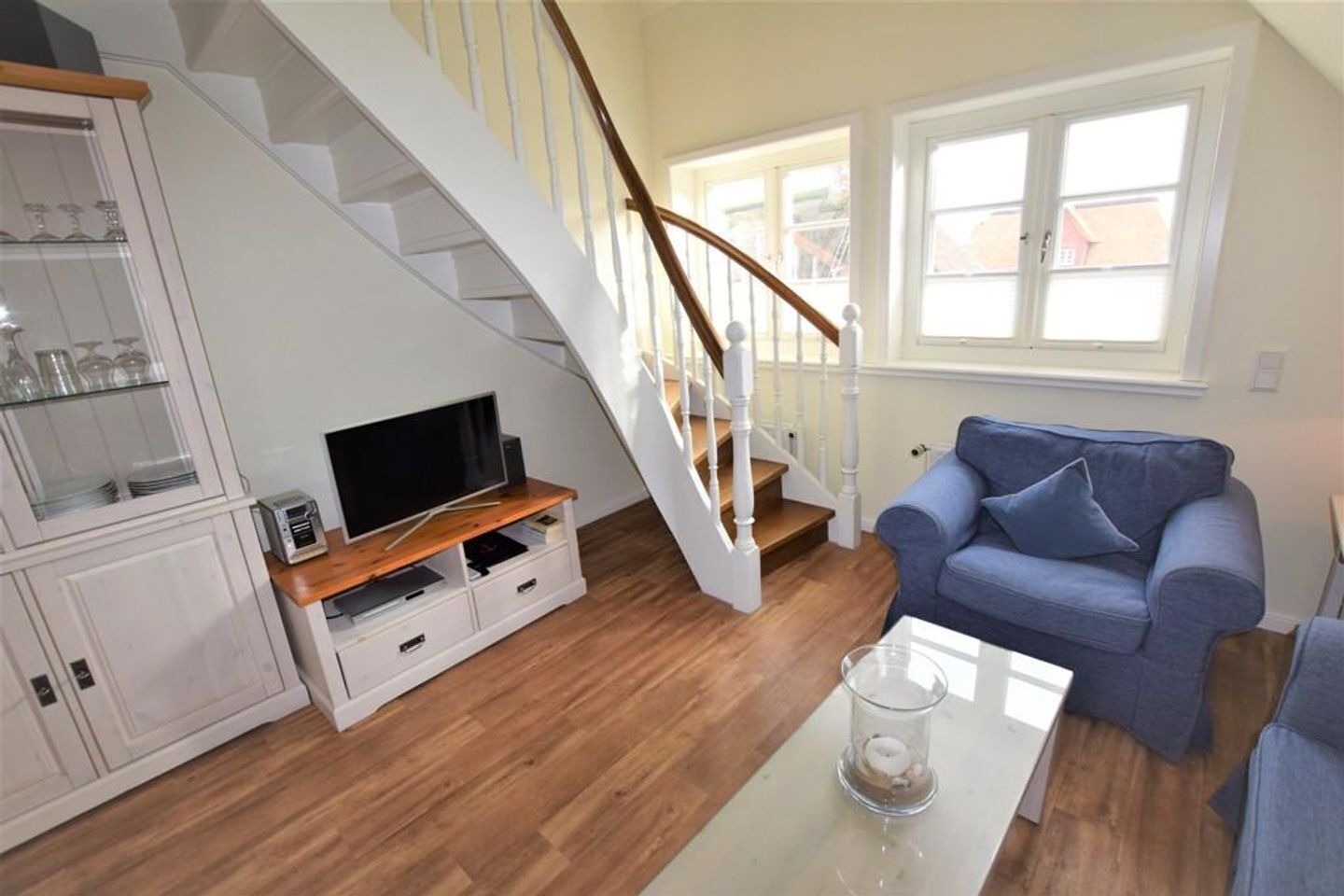 A living room with a staircase, a blue sofa, a glass coffee table, and a TV on a wooden stand.