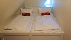 Ein kleines Zimmer, weiß gestrichen, mit zwei Betten, geneigter Decke und einem Fenster für natürliches Licht.