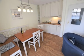 A compact kitchen with white cabinets, a wooden dining table, and a blue armchair. - Landhaus Ruusenhüs App. Ruu 05 - Landhaus Ruusenhüs (Sylt)
