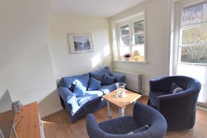 A living room with a blue sofa, a wooden coffee table, and a blue armchair. There is a window with a view of trees and a radiator below it. - Landhaus Ruusenhüs App. Ruu 05 - Landhaus Ruusenhüs (Sylt)