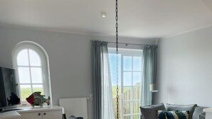 Una sala de estar moderna con un sofá gris, una mesa de comedor de madera con sillas, un televisor en un mueble bajo y un ventanal con cortinas.