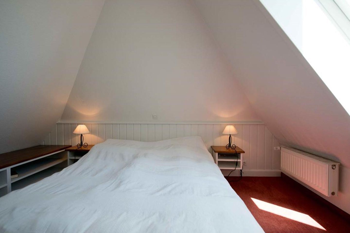 Une chambre avec un plafond en pente, un lit avec une literie blanche, deux tables de chevet avec des lampes et un radiateur fixé au mur.