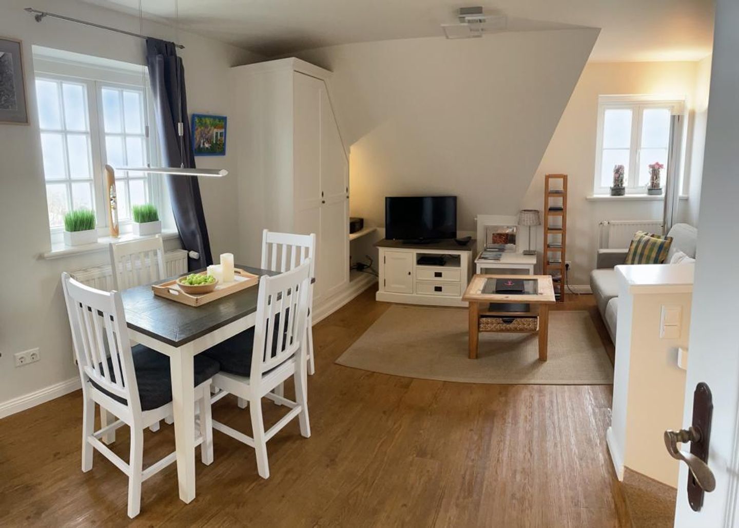 Ein modernes Wohnzimmer mit Essbereich, weißen Möbeln, einem Fernseher und einem Fenster mit Vorhängen.