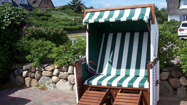 Une chaise de plage en bois, dotée d’un coussin à rayures vertes et blanches, est placée sur une zone pavée, avec un mur de pierre et de la verdure en arrière-plan.