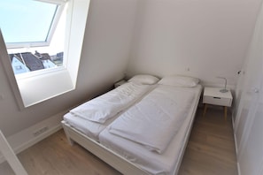 A small, clean room with a single bed, a bedside table, and a window with a view of buildings.