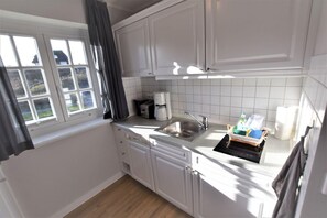 A compact kitchen with white cabinets, a sink, and a window with curtains. - Warfthüs App. 08 - Warfthüs (Sylt)