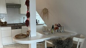 A modern kitchen with white cabinetry, a central island, and a dining area with white chairs. - House Rundblick App. 06 - panoramic view (Sylt)
