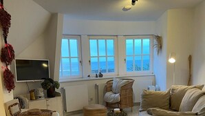 A modern living room with a sofa, a small table, and a TV. There are windows with a view of the sea. - House Rundblick App. 06 - panoramic view (Sylt)