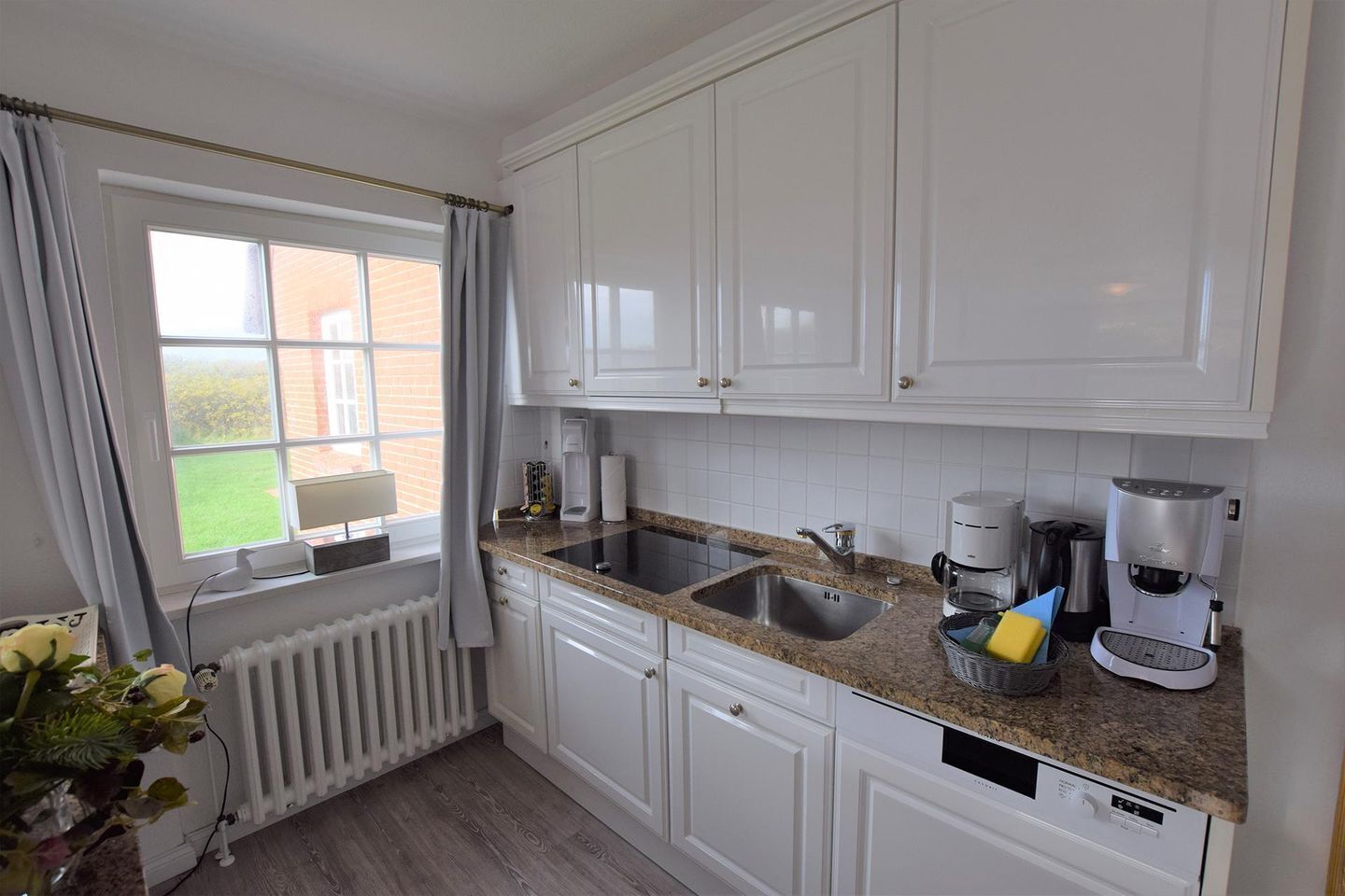 A kitchen with white cabinets, granite countertops, a built-in dishwasher, a coffee maker, and a window with curtains.